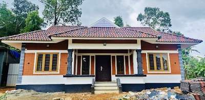 A Blend of Tradition and Innovation at Vazhakulam, Ernakulam

Owner: Sri. Manokumar
Location: Vazhakulam, Ernakulam
Area: 1457 sq.ft
Estimate: ₹25,00,000 + 10% Supervision Charge [excluding interior]
Type: 3 BHK Interlock Home
Designed by: Building Designers – K.V. Muraleedharan

Nestled amidst the lush greenery of Vazhakulam, this 1457 sq.ft interlocking home beautifully bridges the charm of Kerala’s traditional architecture with the sustainability of modern construction. Designed by K.V. Muraleedharan and his expert team at Building Designers, this elegant 3BHK residence is a fine example of cost-effective design and aesthetic excellence.

The exterior, adorned with terracotta-toned interlocking blocks, instantly stands out for its earthy appeal. The tiled sloping roof, with its deep eaves and graceful symmetry, evokes nostalgia for Kerala’s timeless architectural heritage. The front elevation strikes a perfect balance — strong granite-toned pillars enhance the structure’s grandeur, while the warm color palette harmonizes with the natural surroundings.

Inside, the home opens into spacious, well-lit interiors marked by thoughtful detailing. The central pillar section under a coffered wooden ceiling creates a traditional focal point that radiates strength and character. Soft pastel walls complement the wooden ceiling, giving the interiors a serene and inviting aura. Natural ventilation and ample daylight flow through wide windows, ensuring a bright, airy atmosphere throughout the home.

Each of the three bedrooms is designed with functional simplicity, making the most of available space without compromising comfort. The use of interlock technology not only minimizes the need for plastering and painting but also provides superior thermal insulation—keeping the home cool in summer and warm during monsoon months.

Beyond aesthetics, this project is a statement of eco-conscious living and budget-friendly design. Built at a modest cost of ₹25 lakhs (excluding interiors), it demonstrates how sustainable building practices can coexist with refined architectural beauty.

With its traditional soul and modern efficiency, Manokumar’s home at Vazhakulam stands as a proud representation of Building Designers’ vision—homes that are rooted in Kerala’s culture yet crafted for the needs of today’s families.