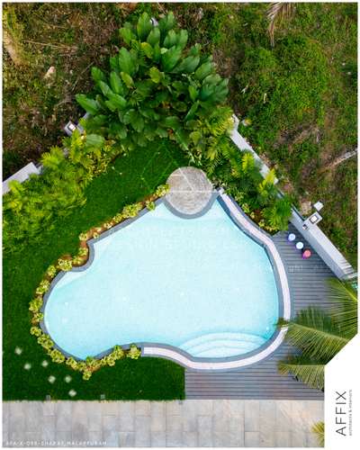 A free-flowing organically-shaped swimming pool with light blue mosaic tiling, featuring built-in steps and a small circular shallow area, all surrounded by a dark gray deck, a border of manicured lawn and low plants, and dense natural foliage on the perimeter #Architect  #architecturedesigns  #Architectural&Interior  #architecturekerala  #kerala_architecture  #best_architect  #swimmingpoolwork